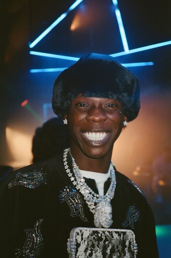 Seyi Vibez smiling in a dark outfit and chain under blue stage lights in London