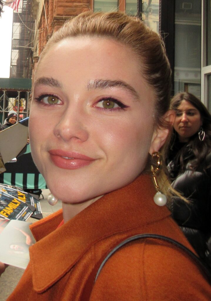 Florence Pugh smiling in an orange coat on a city street